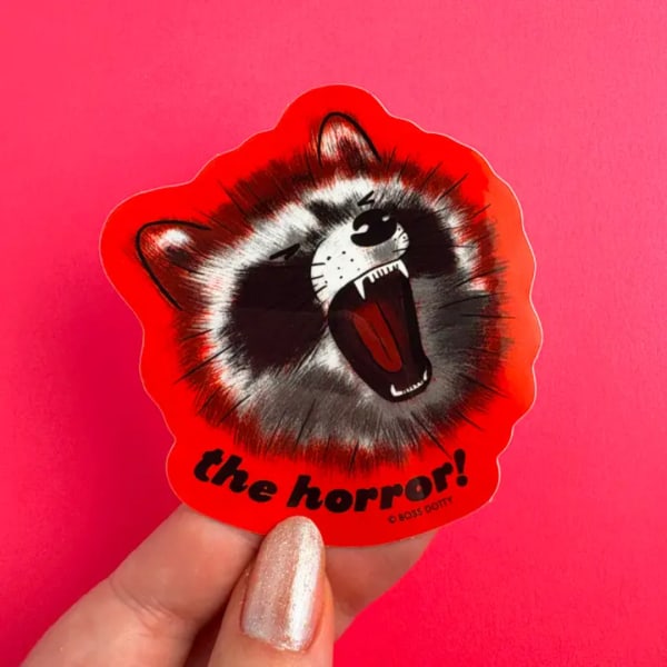 Hand holding a red sticker with a snarling raccoon and the words "the horror!"