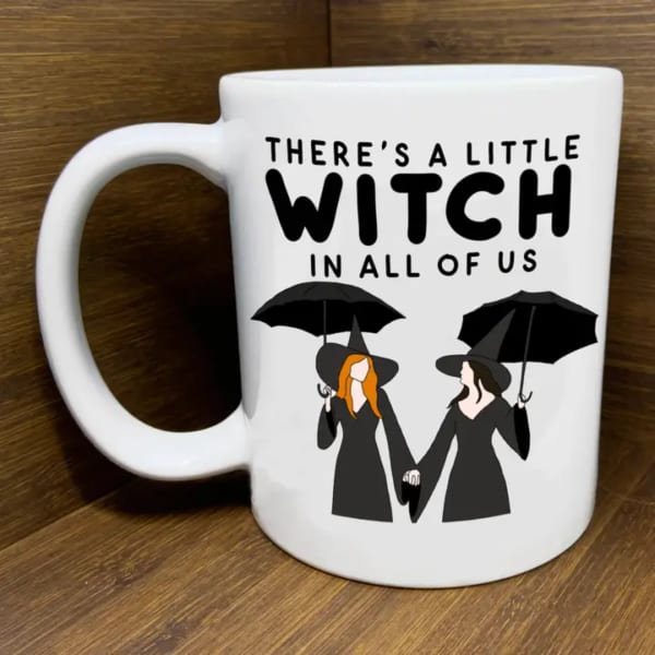 White mug with witch-themed text and two illustrated witches holding umbrellas