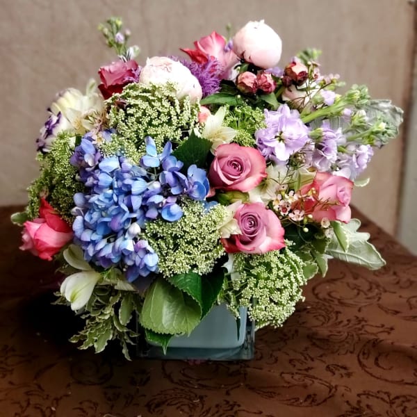 Mixed bouquet of pink, blue, and lavender flowers in a square vase