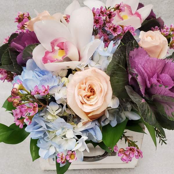 Mixed bouquet with pale roses, orchids, and blue hydrangeas in a white container