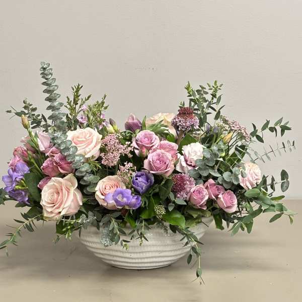 Low centerpiece of pink roses and mixed purple flowers in a white ceramic bowl