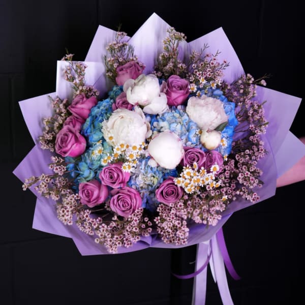 Bouquet of pink roses, blue hydrangeas, and white peonies wrapped in lavender paper