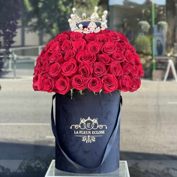 Red rose arrangement in a navy hat box with a jeweled crown topper