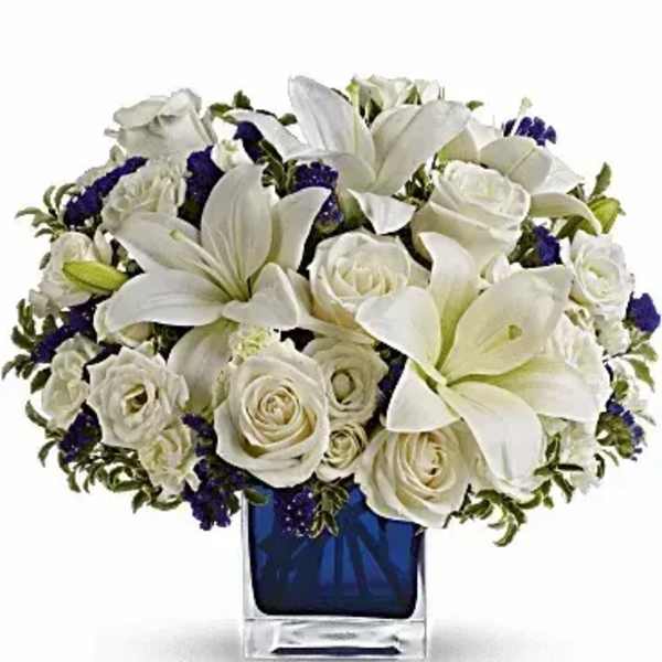 Low arrangement of white lilies and roses with purple accents in a blue cube glass vase