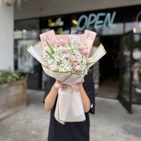 Hand-tied bouquet of light pink tulips and white filler flowers wrapped in pink and white paper with a ribbon.