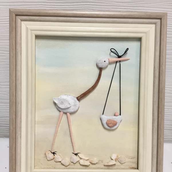 Framed pebble art of a stork carrying a baby in a sling