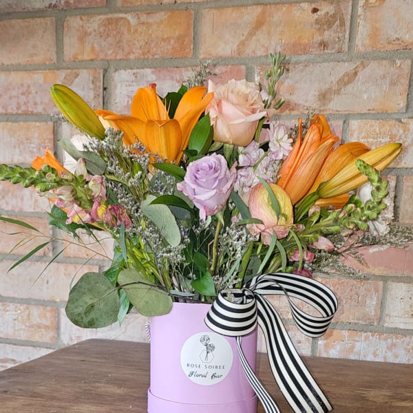 Mixed arrangement of orange lilies and pastel roses in a lavender hatbox with striped ribbon