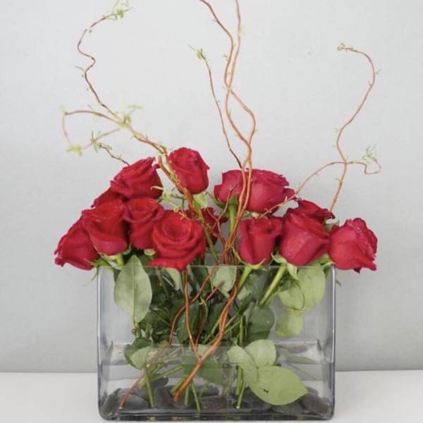 Red roses arranged in a clear rectangular vase with curly branches