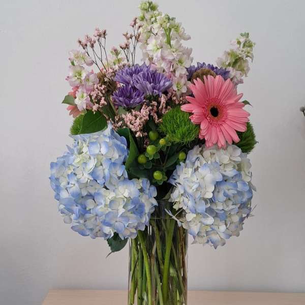 Mixed bouquet with blue hydrangeas, pink gerbera daisy, and purple blooms in a clear vase