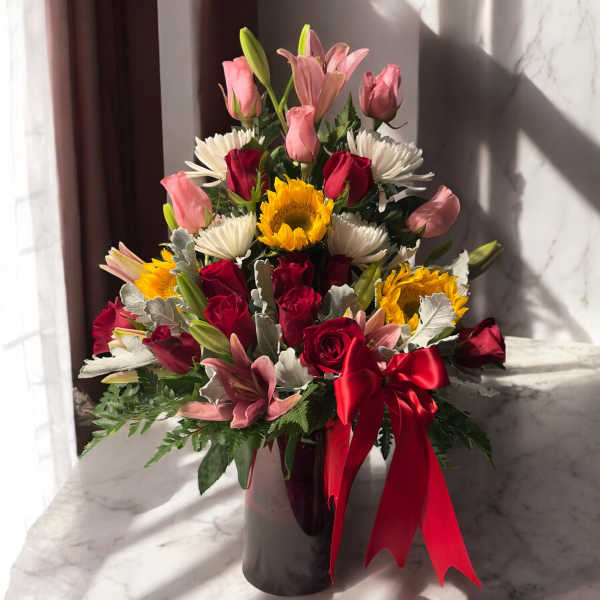 Mixed bouquet of roses, lilies, sunflowers, and white daisies in a vase with a red ribbon