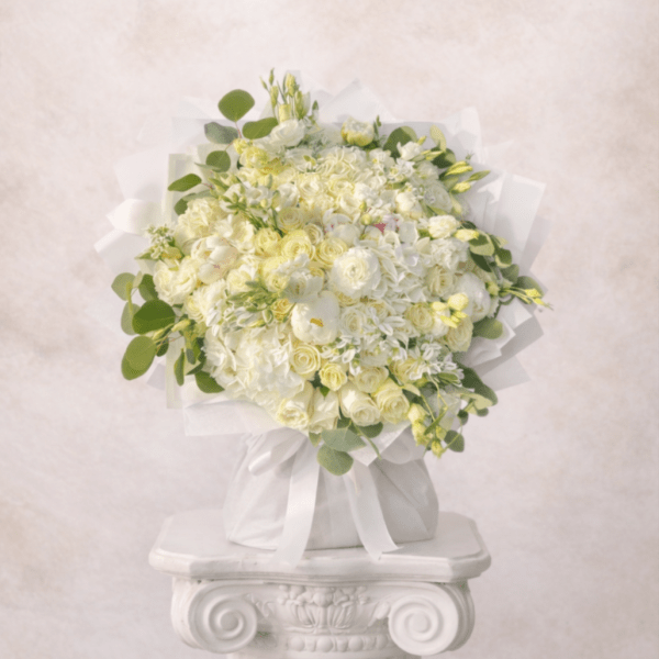 Round bouquet of white and pale yellow flowers wrapped in white fabric on a white pedestal.