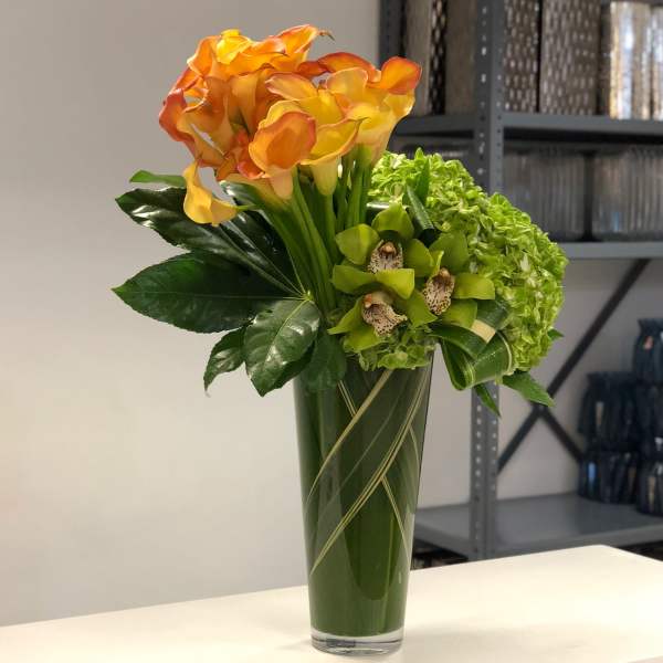 Tall glass vase with orange calla lilies, green hydrangea, and green orchids in a structured arrangement