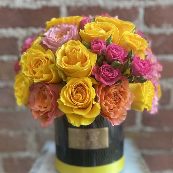 Compact arrangement of yellow, pink, and peach roses in a black hatbox-style container with a yellow band