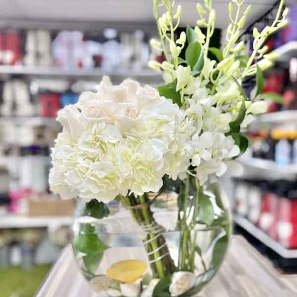Round glass vase of white roses, hydrangeas, and orchids with seashells in the water