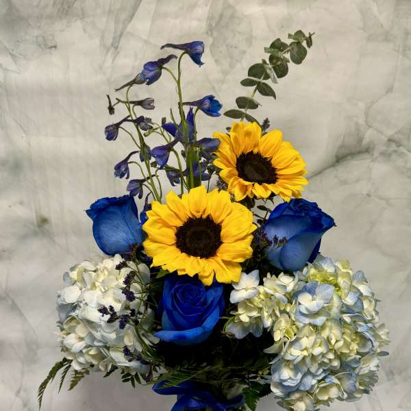 Blue roses and sunflowers in a glass vase with hydrangeas and a ribbon