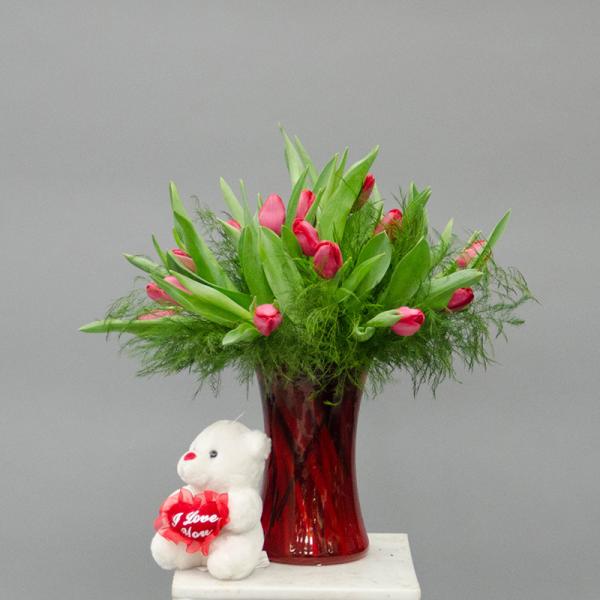 Pink tulips in a red glass vase with a small teddy bear holding a heart