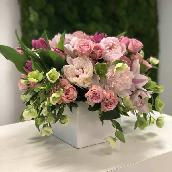 Arrangement of pink roses, peonies, tulips, orchids, and green blooms in a white square vase.