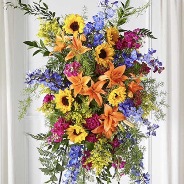 Tall standing spray with sunflowers, orange lilies, blue delphinium, and bright pink blooms on an easel