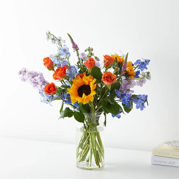 Mixed bouquet with a sunflower, orange roses, and blue blooms in a clear glass vase