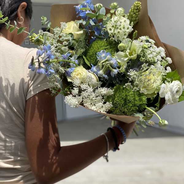 Handheld bouquet of blue, white, and pale yellow flowers wrapped in brown paper