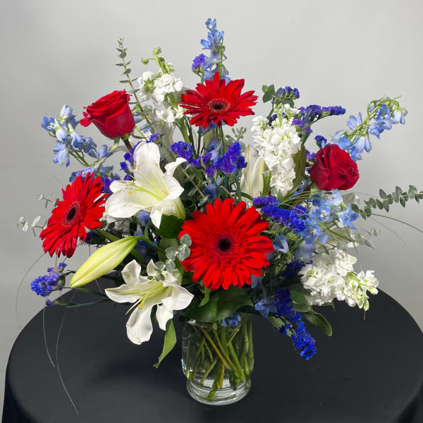Mixed bouquet with red gerbera daisies, red roses, white lilies, and blue flowers in a glass vase
