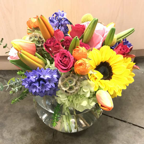 Colorful mixed bouquet in a clear glass vase with sunflowers, roses, tulips, and purple blooms
