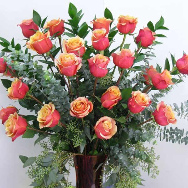 Tall arrangement of pink and yellow roses with lush foliage in a red glass vase