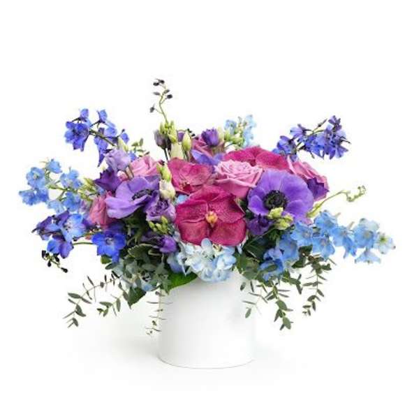 Low arrangement of pink roses and purple and blue blooms in a white cylinder vase