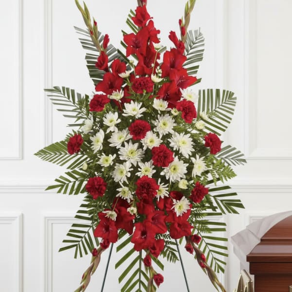 Tall red and white standing spray with carnations and mums on a metal easel beside a wooden casket.