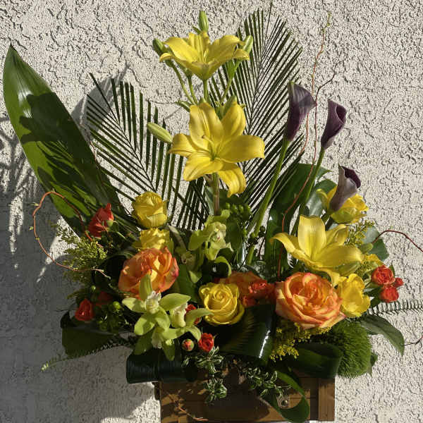 Tall arrangement of yellow lilies, orange and yellow roses, and green orchids in a wooden box