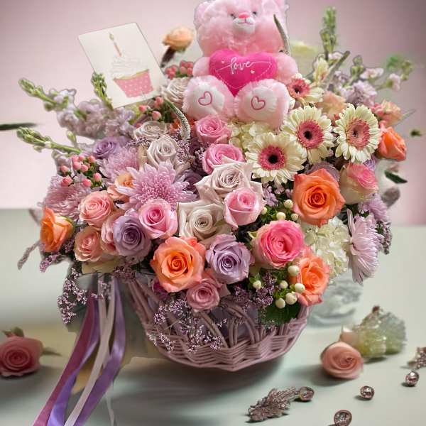 Pink teddy bear in a basket of roses and daisies with a love card
