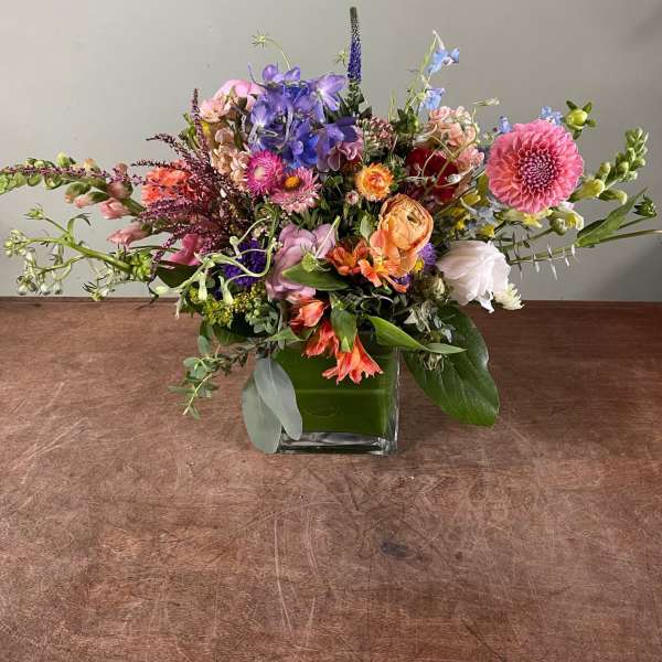 Mixed bouquet in a square glass vase with pink, purple, orange, and white flowers.