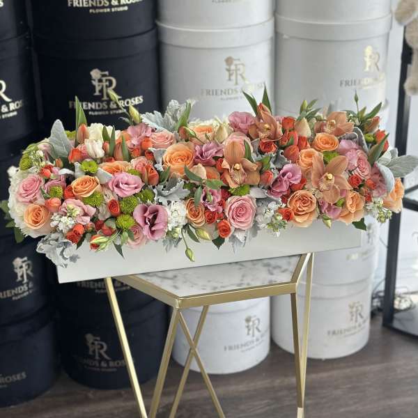 Low rectangular arrangement of pink, peach and orange flowers in a white box on a gold stand