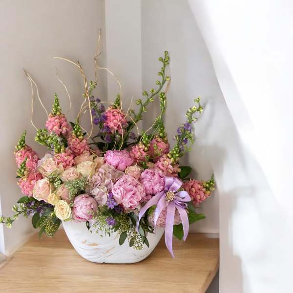 Pink and cream floral arrangement in a white bowl vase with a lavender ribbon