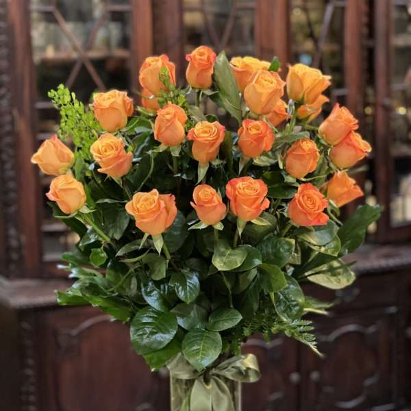 Tall arrangement of orange roses in a clear glass vase with a green ribbon bow.