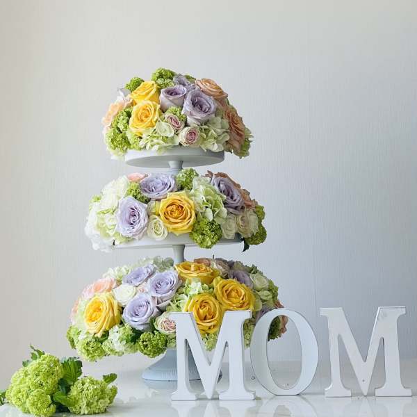 Three-tier floral arrangement with pastel roses and hydrangeas beside large MOM letters