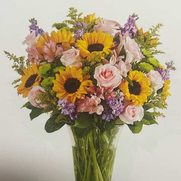 Bouquet of sunflowers, pink roses, and purple flowers in a clear glass vase