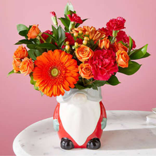 Bright orange and red flower arrangement in a ceramic gnome-shaped vase on a white table.