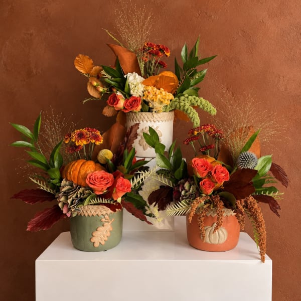 Three autumn floral arrangements in decorative pots on a white pedestal.