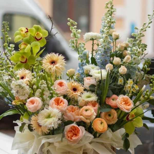 Mixed bouquet in a woven basket with peach, white, blue, and green flowers