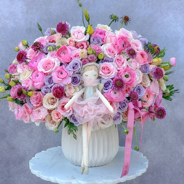 Large pink and lavender rose arrangement with a doll in a white vase