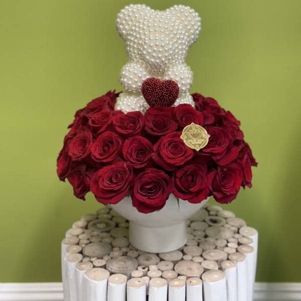 Red roses arranged around a pearl-covered teddy bear in a white vase