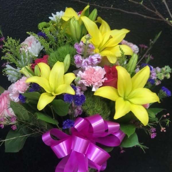 Bouquet of yellow lilies, pink flowers, and a purple ribbon in a glass vase