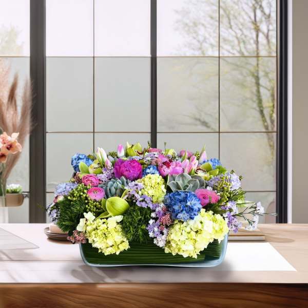Low rectangular arrangement of pink, blue, and yellow flowers in a glass vase