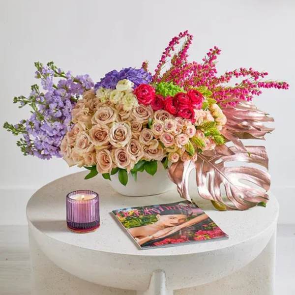 Mixed bouquet of roses and purple flowers in a white vase with a candle and magazine