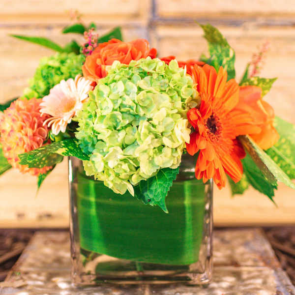 Orange and green floral arrangement in a square glass vase