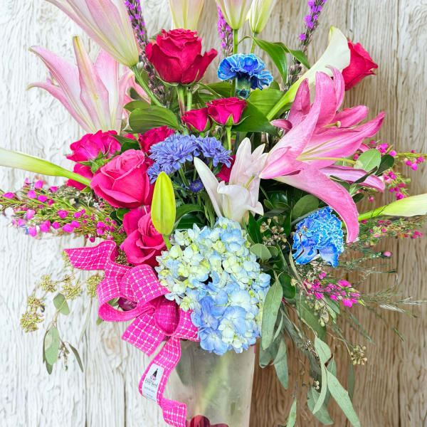 Pink lilies, roses, and blue hydrangeas in a glass vase with a pink ribbon
