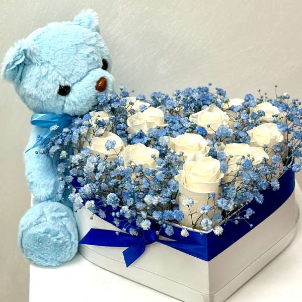 Heart-shaped box of white roses with blue filler flowers and a blue teddy bear plush beside it