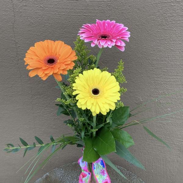 Three colorful gerbera daisies in a glass vase with a floral ribbon