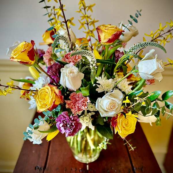 Mixed arrangement of yellow and white roses with pink carnations in a clear glass vase on a wooden table.